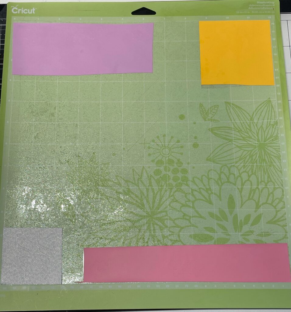 Green Cricut mat with light purple, yellow, silver glitter, and pink vinyl pieces in different corners 
