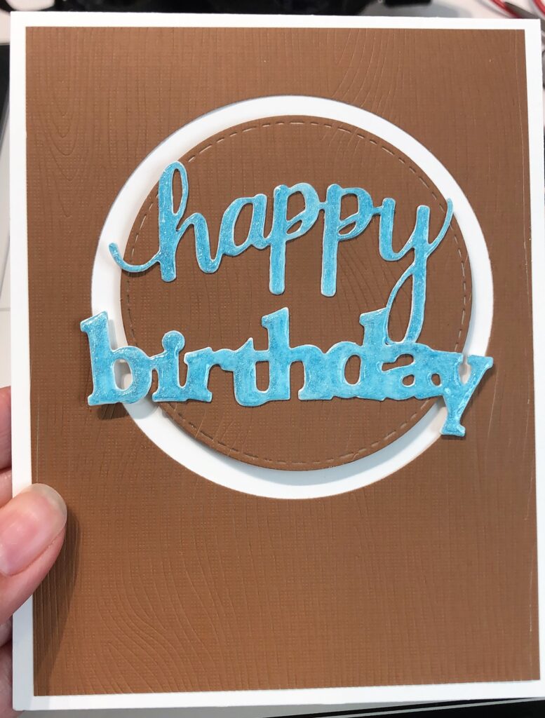 Brown woodgrain birthday card with circle cut out and blue happy birthday words on top of popped up woodgrain circle 