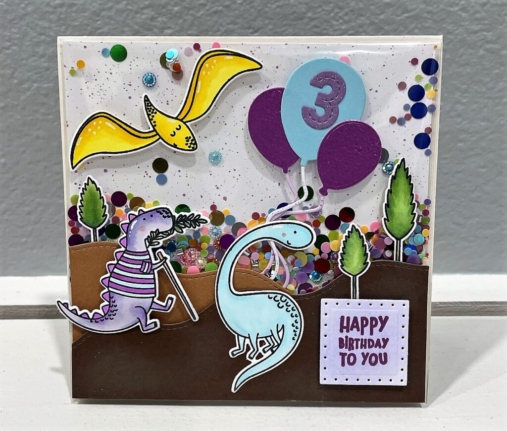 Dinosaur themed shaker birthday card with a light purple dinosaur in a striped purple and blue shirt with a walking stick and a light blue dinosaur holding blue and purple balloons on a mountain scene and yellow flying dinosaur in the top left corner - sentiment in square on bottom right says happy birthday to you 