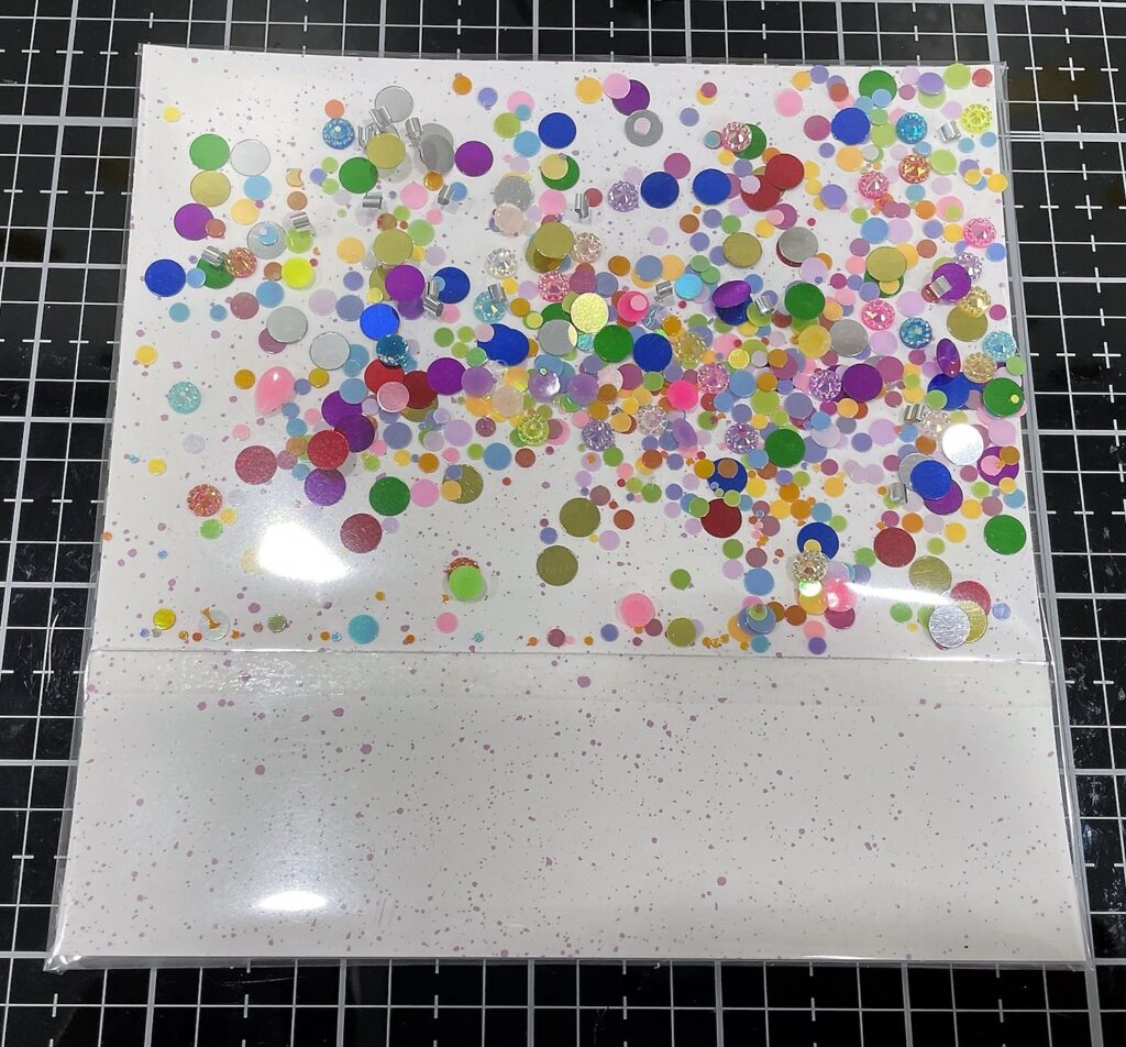 White paper with purple speckled paper with sequins, confetti, and gems inside a thin plastic pocket