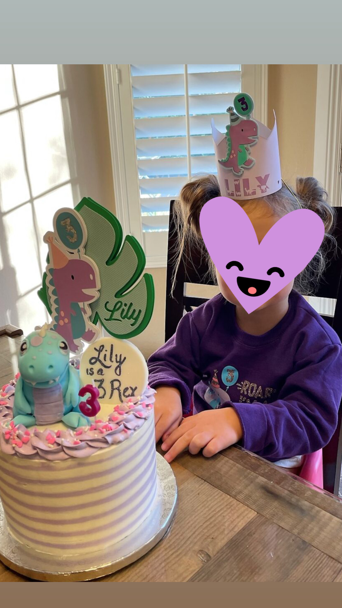 Toddler with purple heart over her face in front of a dinosaur birthday cake