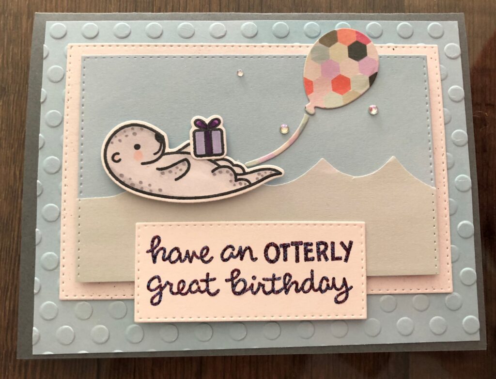Blue and gray birthday card with a light gray otter on its back with a birthday present resting on its belly and a multicolored balloon floating above it and a rectangle with the words "have an otterly great birthday" below the scene