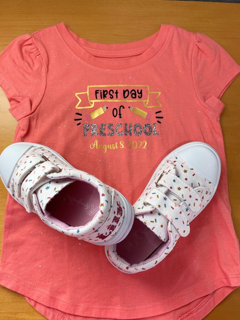Sparkly coral shirt that says "First day of Preschool" and "August 8, 2022" and pencils decorating it. Sitting on top of the shirt are white velcro sneakers with a sprinkle design and sparkly "Lily" on the back heels. 