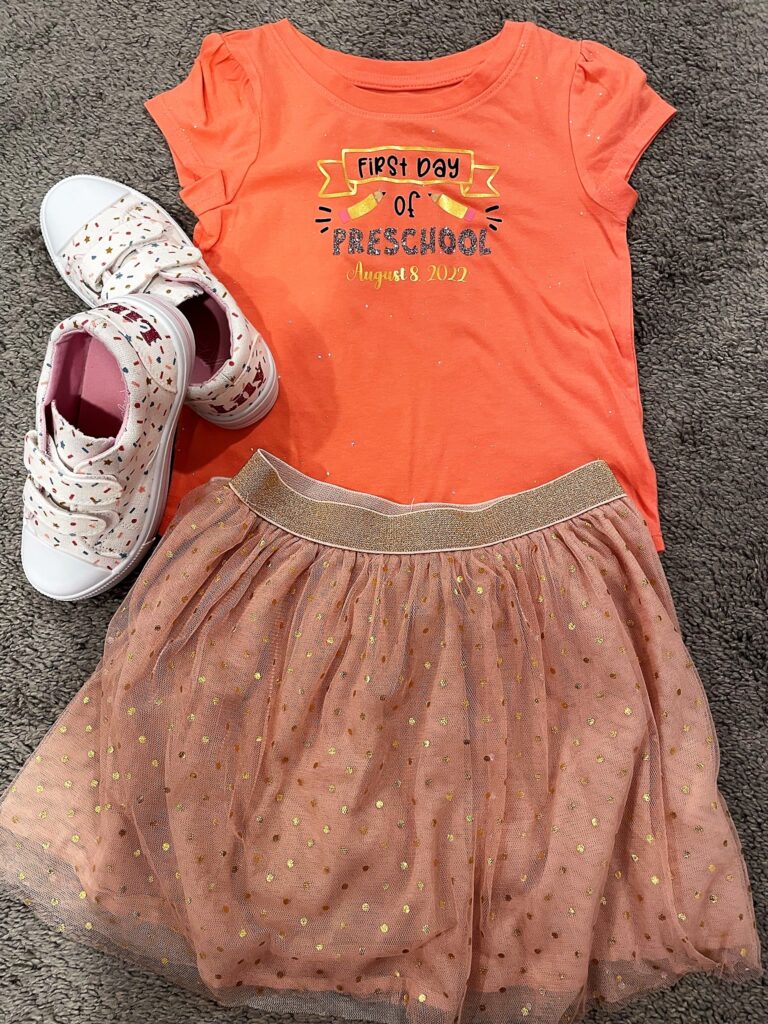 Sparkly coral shirt that says "First day of Preschool" and "August 8, 2022" and pencils decorating it. Sitting on top of the shirt are white velcro sneakers with a sprinkle design and sparkly "Lily" on the back heels and a pink tutu skirt with gold polka dots.